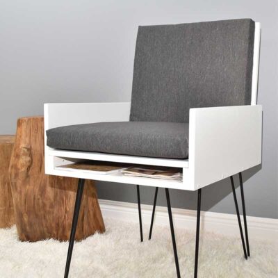 Easy DIY chair with a hidden compartment and book storage, designed with a white frame, gray cushion, and black metal legs on a white rug. A wooden stump side table is positioned to the left. Magazines are stored in the concealed area under the seat. The background wall is light gray.