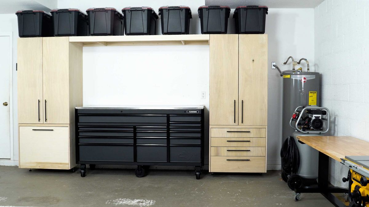 A garage set up for DIY and woodworking includes two tall garage cabinets, multiple storage solutions, a large black rolling tool chest, six black storage bins stacked on top, a visible water heater, a portable air compressor, and a wooden workbench.